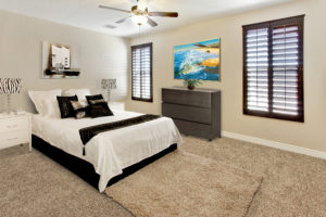 Interior of a Cullers home- bedroom with queen size bed in black and white, beige carpet and two windows with dark wood shutters